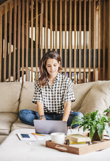 Frau mit langen braunen Haaren sitzt auf einem Sofa mit einem Laptop und einem Notebook und arbeitet. - MINF04626