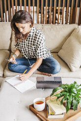 Frau mit langen braunen Haaren sitzt auf einem Sofa mit einem Laptop und einem Notebook und arbeitet. - MINF04629