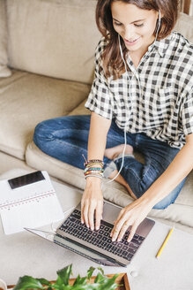 Frau mit langen braunen Haaren sitzt auf einem Sofa mit einem Laptop und einem Notebook und arbeitet. - MINF04630