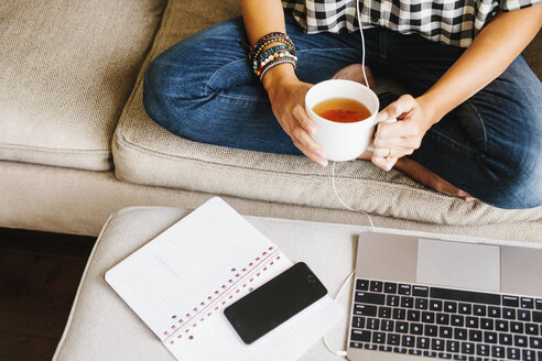 Frau in Jeans sitzt auf einem Sofa vor einem Laptop und hält eine Tasse. - MINF04632