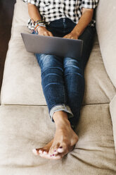 Frau in Jeans, die auf einem Sofa liegt und einen Laptop benutzt. - MINF04633