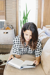 Frau mit langen braunen Haaren liegt auf einem Sofa und liest ein Buch. - MINF04639