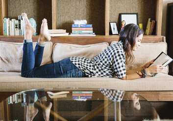 Frau mit langen braunen Haaren liegt auf einem Sofa und liest ein Buch. - MINF04640
