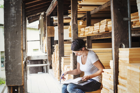 Frau sitzt auf einem Holzlagerplatz und hält einen Ordner. - MINF04675