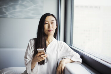Eine Geschäftsfrau, die sich auf die Arbeit vorbereitet, sitzt im Nachthemd am Fenster und hält ein Smartphone in der Hand. - MINF04712