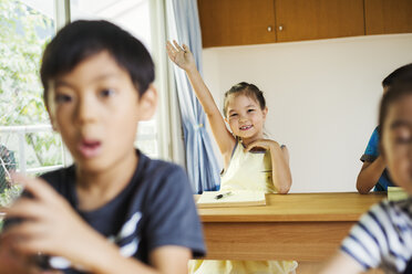 Eine Gruppe von Kindern in einem Klassenzimmer, eines davon mit erhobener Hand, bereit, eine Frage zu beantworten. - MINF04734