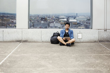 Ein Mann, der auf dem Boden sitzt und sein Smartphone benutzt, vor einem Aussichtsfenster mit Blick auf eine Stadt. - MINF04753