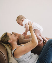 A woman lying on a sofa playing with a baby girl, lifting her in the air. - MINF04772