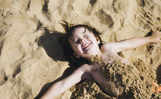 Ein Junge liegt auf dem Rücken am Strand, sein Oberkörper ist mit Sand bedeckt, und lacht. - MINF04789