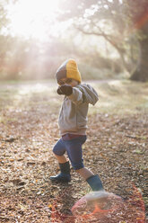 A boy outdoors in a woolly hat wearing shorts and wellington boots, striking a pose with an outstretched arm. - MINF04824