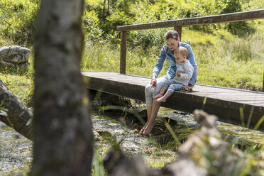 Mother and daughter sitting on wooden bridge, mountain stream - DIGF04724