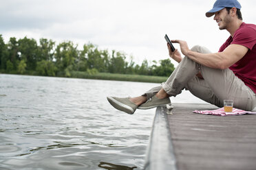 A man seated on a jetty by a lake, using a digital tablet. - MINF04927