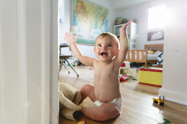 Ein kleines Mädchen, das auf dem Boden eines Schlafzimmers sitzt und lächelnd und mit erhobenen Armen nach oben schaut. - MINF05044