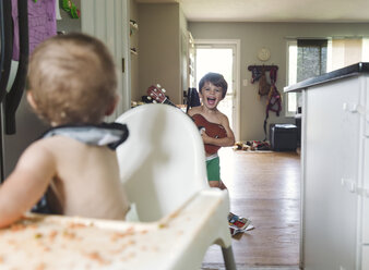 Ein kleines Mädchen sitzt in einem Hochstuhl, der mit verschüttetem Essen bedeckt ist, und blickt hinter sich auf ein Kind, einen Jungen, einen Bruder und eine Schwester. - MINF05050