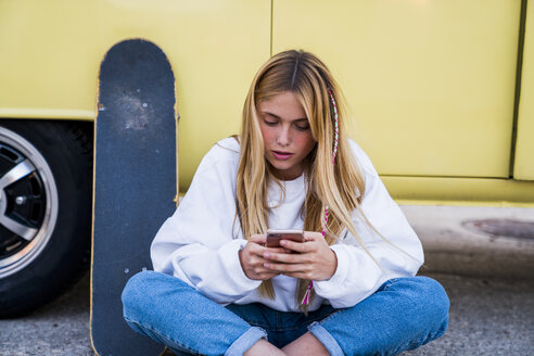 Junge Frau mit Skateboard und Handy sitzt an einem Lieferwagen - KKAF01394