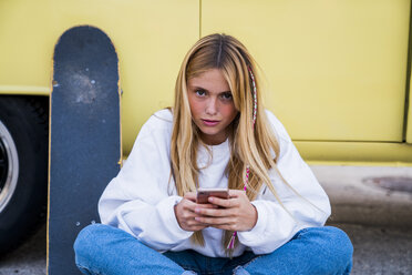Junge Frau mit Skateboard und Handy sitzt an einem Lieferwagen - KKAF01395