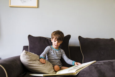 A four year old boy sitting on a sofa at home reading a picture book - MINF05086