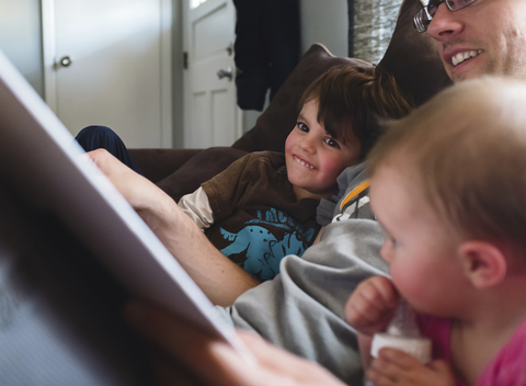 Ein Mann, Vater, liest zwei Kindern, einem Jungen und einem kleinen Mädchen, die auf einem Sofa sitzen, vor., lizenzfreies Stockfoto