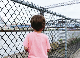 Junge, Kind schaut durch einen Maschendrahtzaun auf einen Fluss. - MINF05096