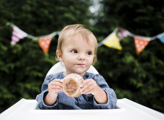 Ein kleines Mädchen sitzt im Hochstuhl im Garten und hält einen Muffin. - MINF05119