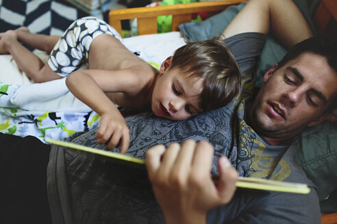 Hoher Blickwinkel auf einen Mann, der im Bett liegt und einem kleinen Jungen mit brauen Haaren eine Gutenachtgeschichte vorliest. - MINF05184