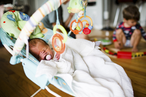 Hoher Blickwinkel auf ein kleines Baby, das in einer Babywippe schläft, ein Junge spielt auf dem Boden im Hintergrund., lizenzfreies Stockfoto