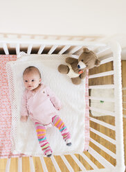 Blick auf ein lächelndes Baby in einem rosa Strampler, das auf dem Rücken in einem weißen Kinderbett liegt, mit einem Teddybär in der Ecke. - MINF05197