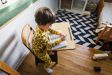 High angle view of young boy with brown hair wearing yellow pyjamas sitting at a table in a nursery, writing into notebook. - MINF05200