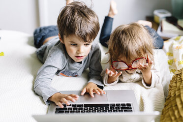 Hoher Blickwinkel auf einen Jungen und ein Mädchen, die eine Brille für Erwachsene tragen, auf dem Kopf stehend, auf einem Bett liegend, mit einem Laptop. - MINF05212