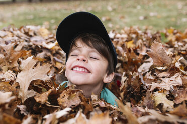 Close up of smiling young boy wearing baseball cap sitting in a heap of autumn leaves. - MINF05238