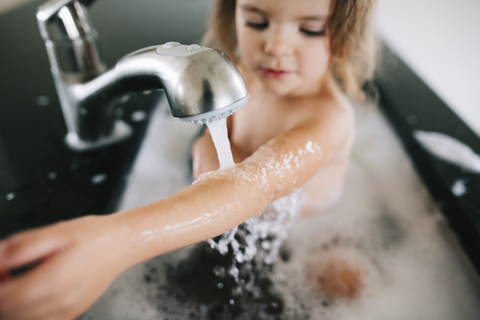 Blick von oben auf ein junges Mädchen, das in einer Badewanne sitzt und ihren Arm unter fließendes Wasser aus einem Wasserhahn hält., lizenzfreies Stockfoto