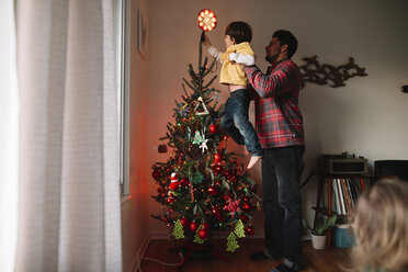 Ein Mann hält einen kleinen Jungen hoch, der einen beleuchteten Weihnachtsbaum schmückt. - MINF05244