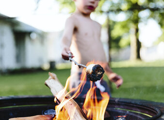 Kind, Junge, der in einem Garten steht und Marshmallows auf einem Stock in einer Feuerstelle röstet. - MINF05252