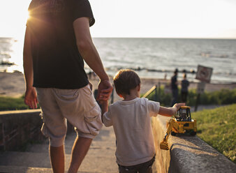 Rückansicht einer Frau und eines Jungen, die sich an den Händen halten und eine Treppe am Meer hinuntergehen, der Junge hält einen Spielzeugbagger. - MINF05272