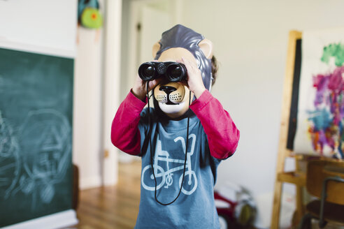 Young boy wearing lion mask standing indoors, looking through a pair of binoculars. - MINF05278