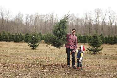 Ein Junge und ein Mann halten eine Säge und einen Weihnachtsbaum in der Hand und stehen im Freien in der Nähe eines Waldes. - MINF05280