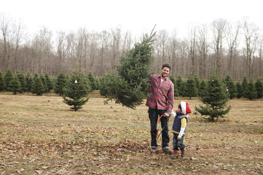 Young boy and man holding saw and Christmas tree standing outdoors near a forest. - MINF05280