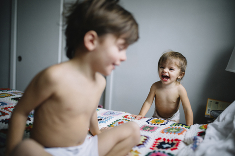 Junger Junge mit nacktem Oberkörper auf einem Bett sitzend, lächelndes junges Mädchen in Windel neben dem Bett stehend., lizenzfreies Stockfoto