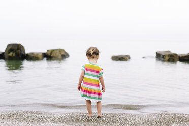 Rückansicht eines jungen Mädchens im gestreiften Sommerkleid, das an einem Sandstrand steht und das Meer beobachtet. - MINF05297