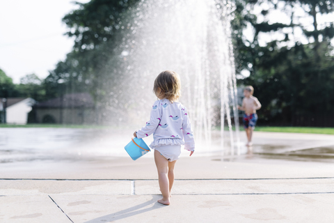 Rückansicht eines jungen Mädchens, das an einem Wasserbrunnen steht und einen blauen Eimer hält., lizenzfreies Stockfoto