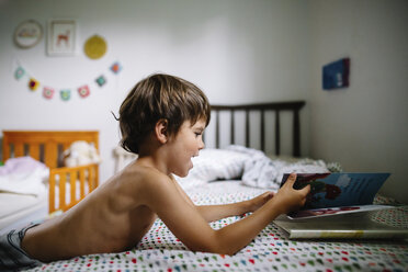 Junger Junge mit nacktem Oberkörper und braunem Haar liegt auf dem Rücken auf einem Bett in einem Kinderzimmer und betrachtet ein Buch. - MINF05306
