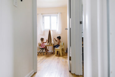 Blick durch eine offene Tür in ein Kinderzimmer, ein Junge und ein Mädchen sitzen auf gegenüberliegenden Seiten einer Staffelei und zeichnen. - MINF05318