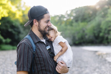 Smiling bearded man wearing baseball cap standing outdoors, holding young girl. - MINF05325
