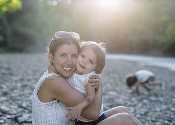 Smiling woman and young girl with brown hair sitting on a pebble beach by a stream, hugging. - MINF05326