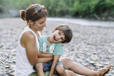 Smiling woman and young boy with brown hair sitting on a pebble beach by a stream. - MINF05327