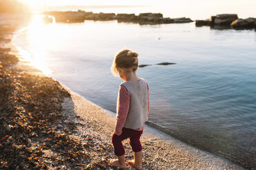 Rückansicht eines jungen Mädchens, das an einem Sandstrand am Meer spazieren geht, Sonnenuntergang. - MINF05339
