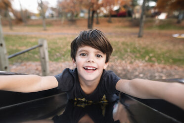 Portrait of young boy with brown hair sitting on a slide on a playground, smiling at camera. - MINF05340