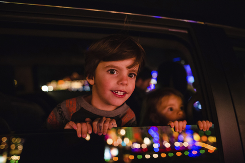 Junge und Mädchen sitzen in einem Auto und schauen aus dem Fenster, auf dem Glas spiegelt sich der beleuchtete Weihnachtsschmuck., lizenzfreies Stockfoto