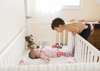 Junge mit braunem Haar, der sich über ein Baby in einem rosa Strampler beugt, das auf dem Rücken in einem Kinderbett liegt. - MINF05385