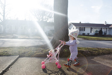 Junges Mädchen mit Strampler und Strickmütze, das einen Spielzeugkinderwagen auf einem Bürgersteig schiebt. - MINF05399